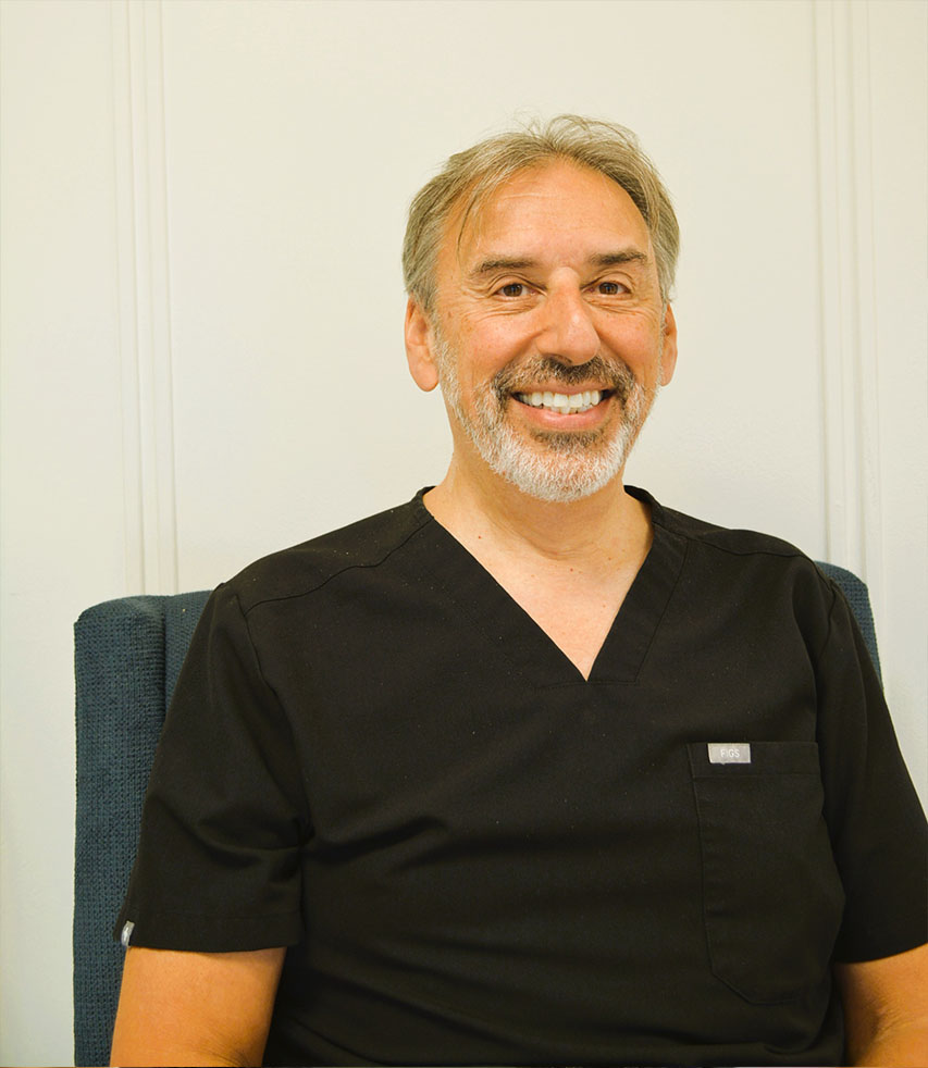 The image shows a man with gray hair, wearing glasses, sitting in a chair with a white wall behind him. He has a beard and mustache, and he is smiling at the camera.