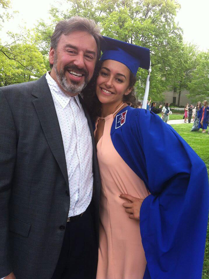 A man and woman posing together at an outdoor event, with the man wearing a suit and tie and the woman in graduation attire, including a blue gown and cap.