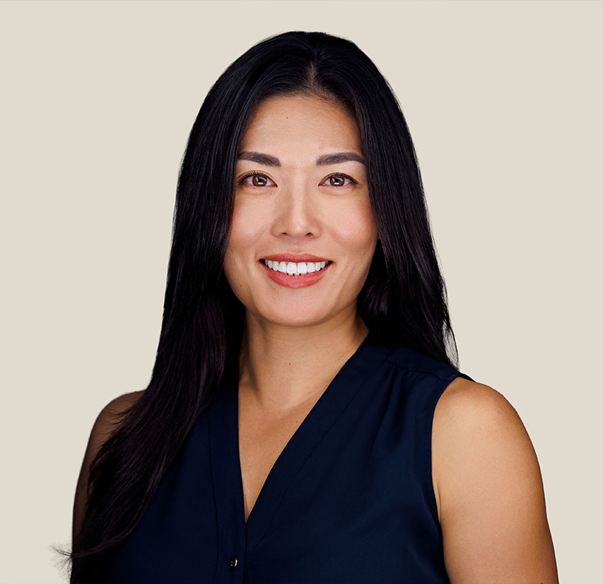 A woman with long dark hair smiling at the camera, wearing a green dress, standing indoors with a modern office environment in the background.