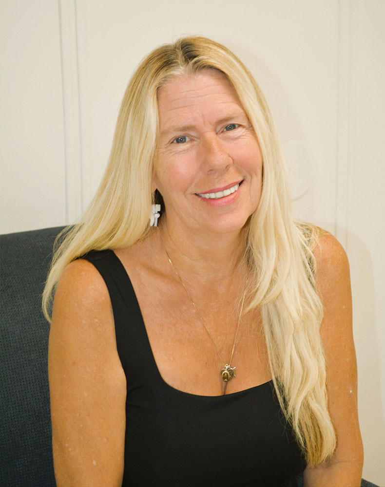 The image features a woman with blonde hair smiling at the camera, wearing a black top, sitting indoors with a blurred background.