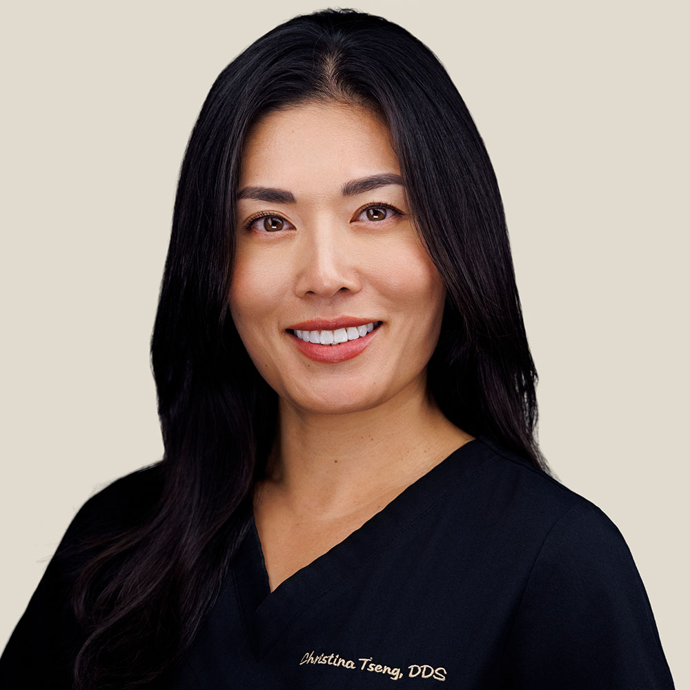 The image shows a smiling woman with dark hair, wearing a black top with a name tag, posing for a photo with a blurred background.