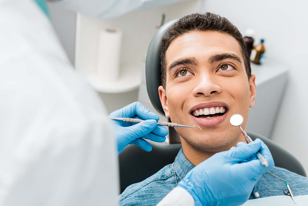 An adult male with a big smile sitting in a dental chair, receiving dental treatment from a dentist who is adjusting his teeth.