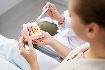 The image depicts a dental professional holding a model of a human mouth with teeth, using a magnifying glass to inspect or demonstrate it, while another individual watches attentively from behind.